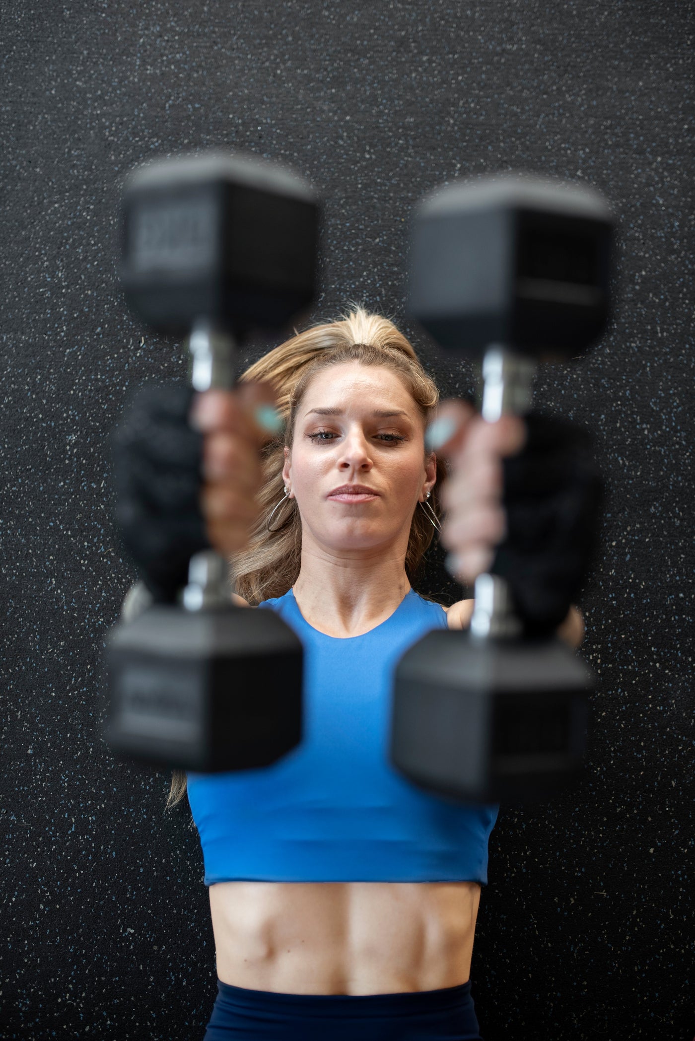 Top view of a woman lying on a gym floor mat wearing a blue cropped gym shirt and lifting and exercising with dumbbells.