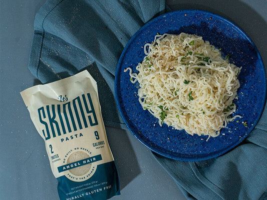 A blue plate of angel hair pasta, topped with herbs, laying next to a pouch of It's Skinny Angel Hair Pasta. 