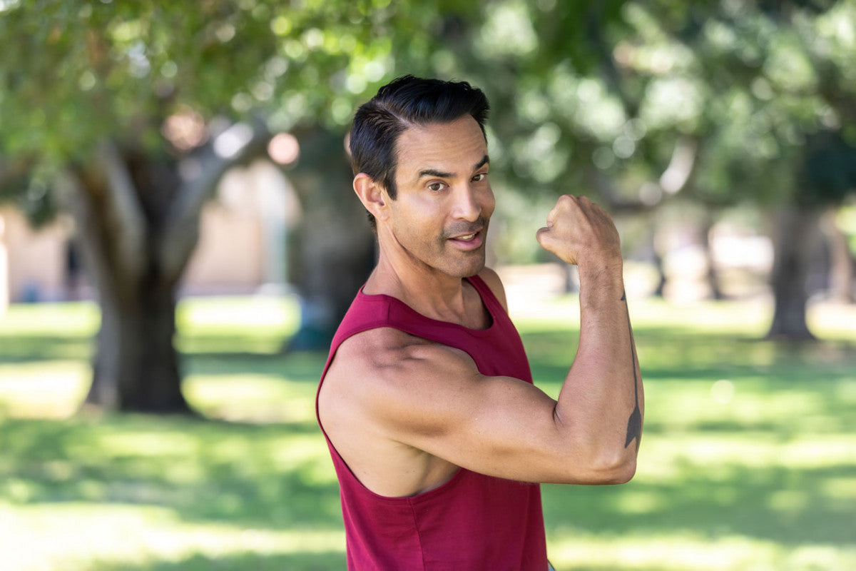 An image of Jorge Cruise in a maroon workout tank in the park, showcasing his biceps and triceps.
