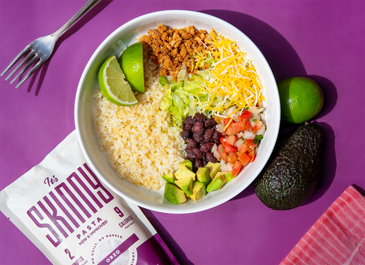 An overhead image of a low-carb taco rice bowl on a purple background. The taco bowl is garnished with lime wedges, diced avocado, pico de gallo, and shredded cheese.