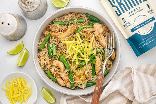 A bowl of yummy chicken, ginger chicken pasta, topped with a dairy free cheese option and surrounded by lime wedges, salt and pepper shakers and a package of angel hair It's Skinny Pasta