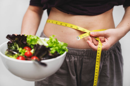 The mid-section of a female is shown with belly visible and being measured with yellow measuring tape. The individual is holding a white porcelain bowl with a mixed, spring salad and cherry tomatoes.