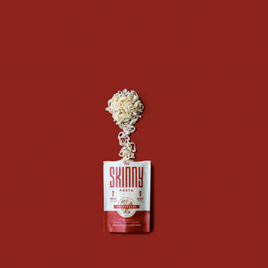 A bag of It's Skinny Fettuccine pasta on a red background, with flat Fettuccine noodles flowing from the open top.